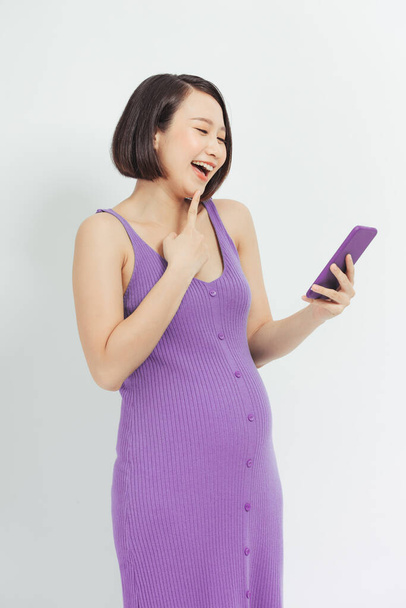 Mujer embarazada emocionada viendo contenido multimedia en línea en un teléfono móvil sobre fondo blanco - Foto, imagen