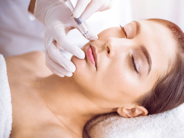 Beautician doing beauty procedure with syringe to face of young brunette woman in sunny spa center. Cosmetic medicine and surgery, beauty injections - Foto, Imagem