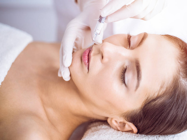 Beautician doing beauty procedure with syringe to face of young brunette woman in sunny spa center. Cosmetic medicine and surgery, beauty injections - Valokuva, kuva