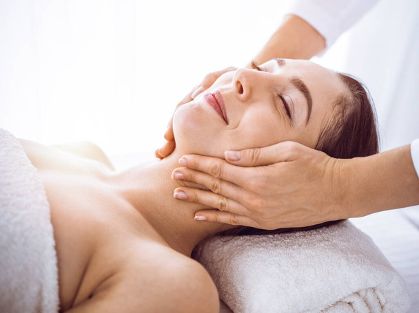 Beautiful brunette woman enjoying facial massage with closed eyes in sunny spa center . Relaxing treatment and cosmetic medicine concepts - Foto, Imagen