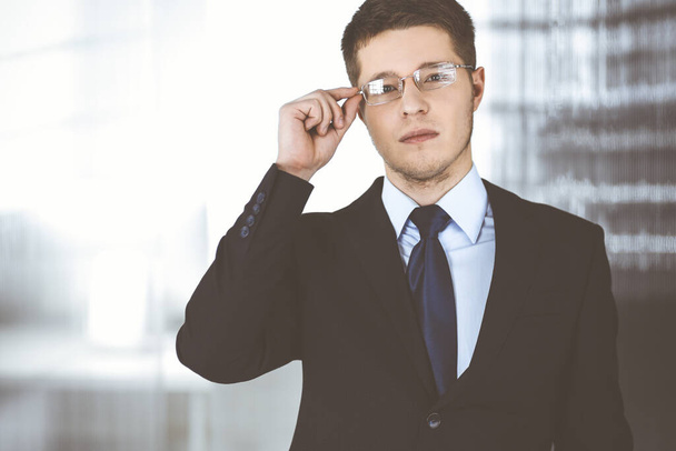 Young businessman wearing suit, with glasses, while standing in the office. Business success concept nowadays - Foto, afbeelding