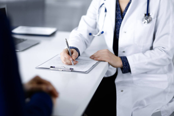 Unknown woman-doctor is writing some medical recommendations to her patient, while they are sitting together at the desk in the cabinet in a clinic. Physician is using a clipboard, close-up. Perfect medical service in a hospital. Medicine concept. - Photo, image