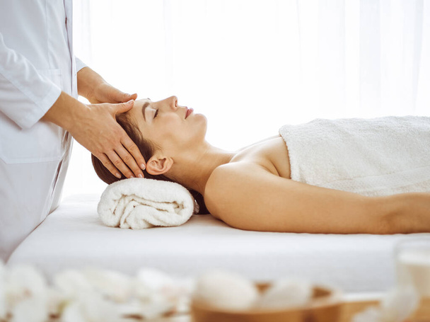 Beautiful brunette woman enjoying facial massage with closed eyes. Relaxing treatment in medicine and spa center concepts - Photo, image