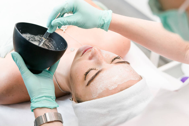 The young female client of cosmetic salon having a cleansing facial mask. The procedure of applying a peeling mask to the face. Concepts of skin care and beauty salon or clinic. - Photo, Image
