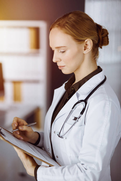 Cheerful smiling female doctor using clipboard in sunny clinic. Portrait of friendly physician woman at work. Medicine concept - Photo, Image