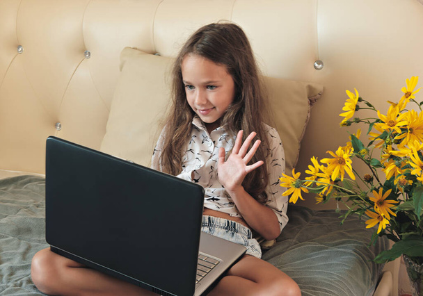 Ein Mädchen kommuniziert mit einem Freund online per Videoanruf auf einem Laptop. Schülerin mit Videokonferenz-App. Videochat-Konzept. Ein Mädchen sitzt auf einem Bett und winkt einem Lehrer zu. Online-Bildung. - Foto, Bild