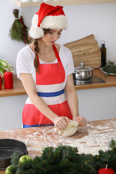 Young brunette woman cooking pizza or handmade pasta while wearing Santa Claus cap in the kitchen. Housewife preparing dough on wooden table - Фото, изображение