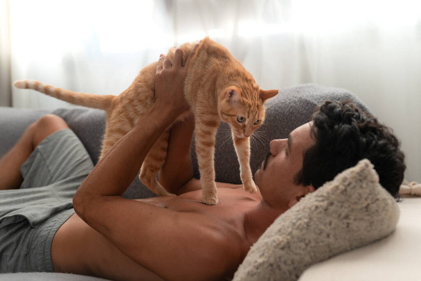 Kanepede yatan genç adam kahverengi bir tekir kediyle oynuyor. - Fotoğraf, Görsel
