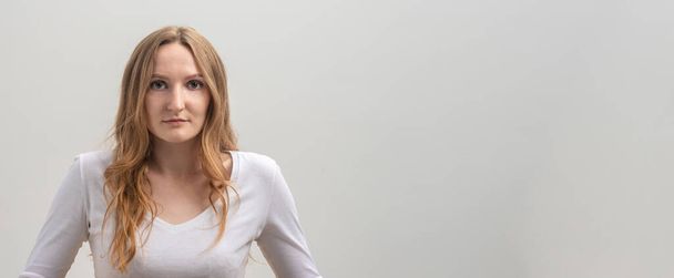 Studio portrait of serious focused caucasian young woman wearing white T-shirt, looking calmly and attentively at camera, over gray background, long golden hair, copy space, banner - Photo, image