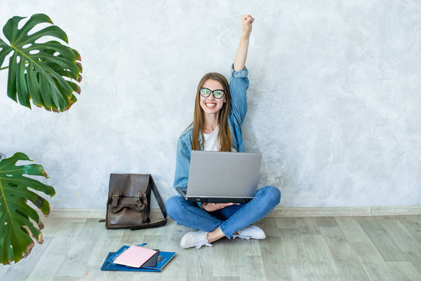 Szczęśliwa młoda studentka w okularach siedzi na podłodze nogi skrzyżowane z laptopem, świętując sukces, podnosząc rękę na szarym tle.Kobieta w dżinsowych ubraniach.Freelancer wykończenie pracy - Zdjęcie, obraz