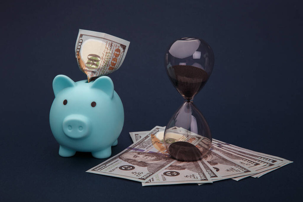 青い背景にお金の銀行券と砂時計と青い貯金箱。時間とはお金の概念 - 写真・画像