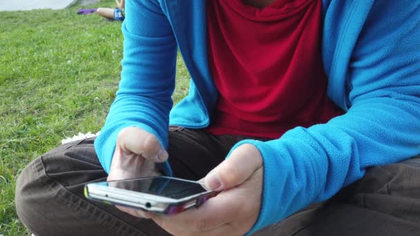 a young womans hands as she looks at her mobile phone sitting on a lawn - Footage, Video