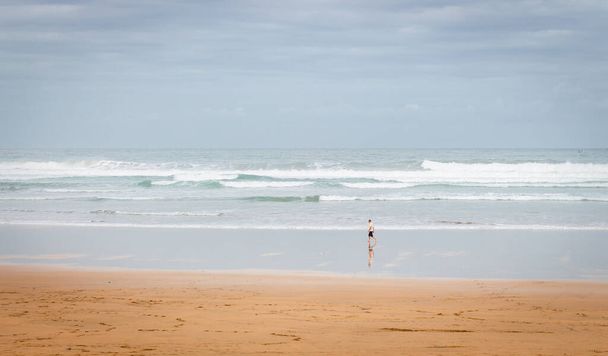 El hombre solitario camina a orillas del mar en un día nublado. Enorme ola en la playa con turista. Paisaje marino panorámico. Otoño en la costa, España. Ilustración de vacaciones. Concepto de deporte surf. Naturaleza costera, viñeta.  - Foto, Imagen