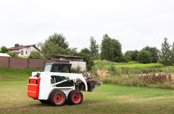 Ein Kompaktlader räumt die Baustelle frei. Landarbeit durch Gebietsverbesserung. Maschine für die Arbeit in beengten Bereichen. Kleintraktor mit Eimer zum Bewegen von Boden, Rasen und Schüttgütern. - Foto, Bild