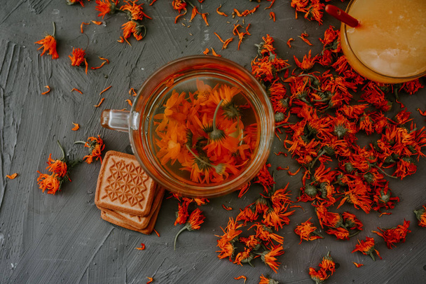 Tea with calendula flowers and biscuits. Transparent glass cup and saucer. Medicinal herbal dried plants marigold, orange calendula. Neutral white and gray background. - Fotografie, Obrázek