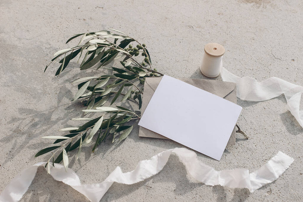 Summer wedding stationery mock-up scene. Blank greeting card, envelope, silk ribbon and olive branches and fruit in sunlight. Grunge concrete background with shadow. Feminine flat lay, top view. - 写真・画像