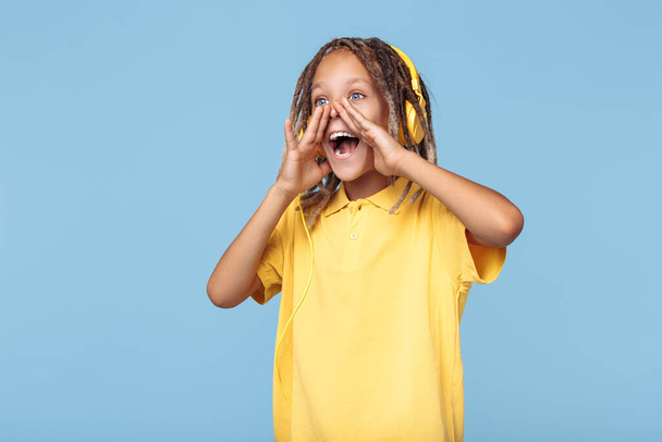 Funny little boy with african dreads listening music in headphones screaming over blue background. - Photo, image