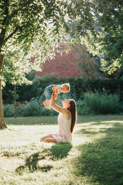 Junge lächelnde kaukasische Mutter und ihr kleiner Sohn spielen im Park. Familienmutter und Kind umarmen sich und amüsieren sich an einem Sommertag im Freien. Glücklicher authentischer Lebensstil in der Kindheit.  - Foto, Bild