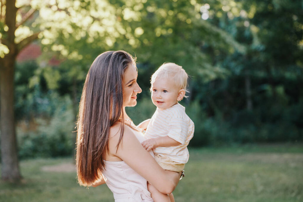 Young smiling Caucasian mother and boy toddler son hugging in park. Mom embracing child baby on summer day outdoor. Happy authentic family childhood lifestyle.  - Photo, Image