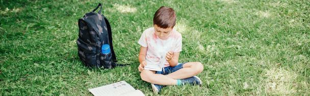Elementary school boy assis dans le parc en plein air faire des devoirs scolaires. Un enfant lit un livre dehors. Auto-apprentissage étudier. Développement précoce pour les enfants. En-tête de bannière Web. - Photo, image