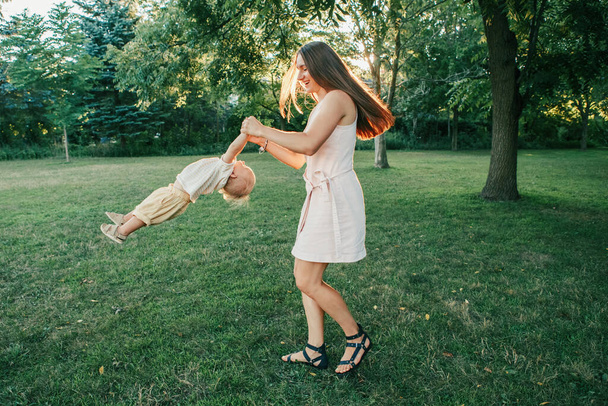 Young mother spinning toddler baby boy outdoor. Parent playing with child son in park. Authentic lifestyle funny moment. Happy family life concept. Childhood and motherhood. - Foto, Bild