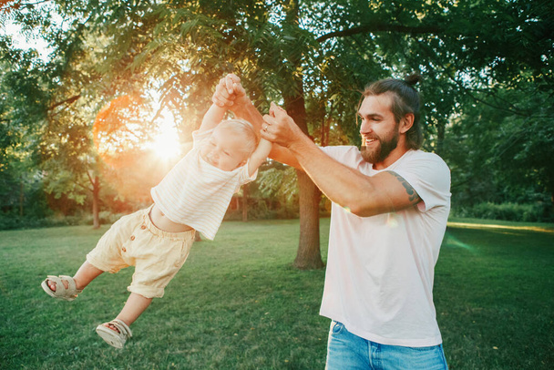 誇り高い父は幼児の赤ちゃんの男の子を屋外に回転させる。公園で子供の息子と遊ぶ親。本物のライフスタイル面白い瞬間。幸せなパパの家族生活の概念。幼少期・父親. - 写真・画像