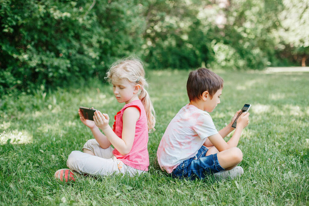 Boy and girl play games on smartphones outdoor. Kids digital gadget screen addiction. Children friends playing online sitting on grass in park. Contemporary problem of loneliness together. - Foto, Imagem