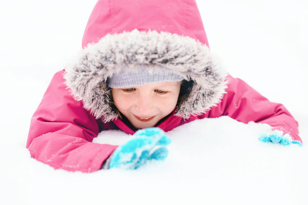 Garota sorridente branca engraçada em roupas quentes de inverno jaqueta rosa brincando com a neve. Criança bonito deitado no chão durante o inverno frio dia nevado na neve. Crianças atividade sazonal ao ar livre. - Foto, Imagem