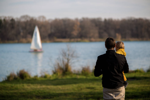 Dad holds his son in a yellow jacket in his arms, they stand with their backs to the camera and look at the lake. Autumn park, Indian summer. - Foto, immagini