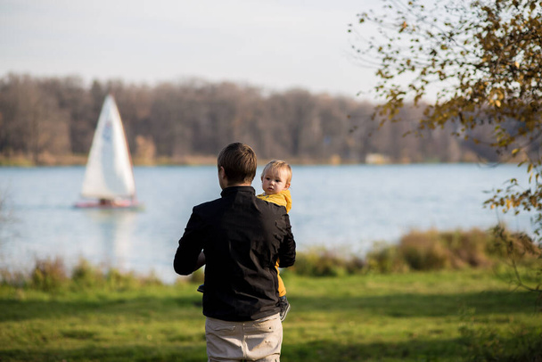 Dad is holding his son in a yellow jacket in his arms, dad is standing with his back to the camera son is looking at the camera against the background of the lake. Autumn park, Indian summer. - Photo, Image