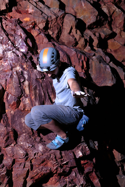 Confident hispanic male climber with his climbing gear on, looking down and waving as he rests on a steep intense red iron rock wall. - Photo, Image