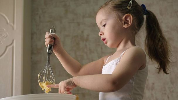 Ein kleines Mädchen probiert rohen Teig mit Schneebesen und Peitschen. Kind bereitet Frühstück zu. Flüssiger hausgemachter Teig für Pfannkuchen und Pfannkuchen. Gesundes Frühstück für das Baby - Foto, Bild