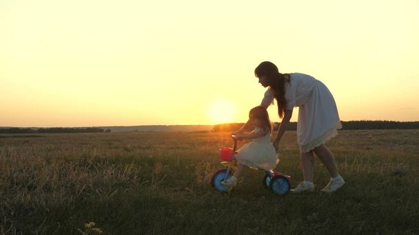 Anne küçük bir çocuğa gün batımında parkta bisiklet sürmeyi öğretiyor. Takım çalışması. Siluet aile yürüyüşü - Fotoğraf, Görsel