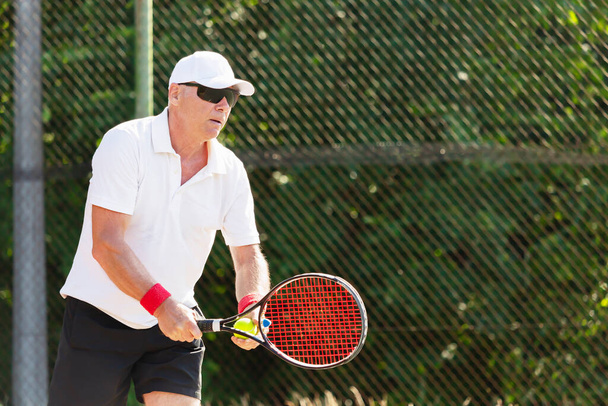 Ein älterer Tennisspieler in guter körperlicher Verfassung spielt an einem sonnigen Tag Tennis auf einem Außenplatz - Foto, Bild