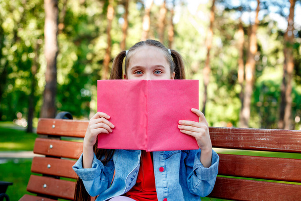Image Of A Little Girl Sitting Outdoors In Nature Park Covering Face With Book. - Photo, Image