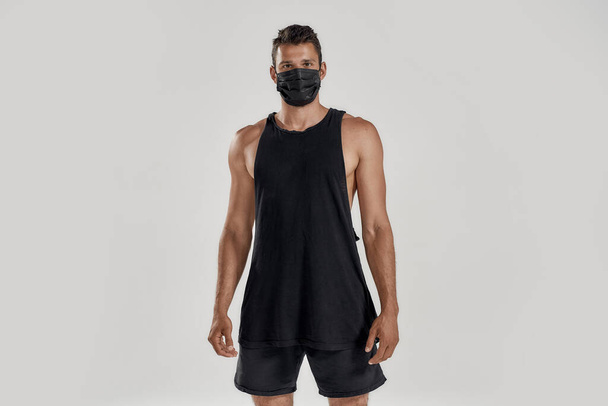 Protection. Portrait of young muscular caucasian man wearing black medical mask looking at camera while standing isolated over grey background. Health, sports, quarantine concept - Fotoğraf, Görsel