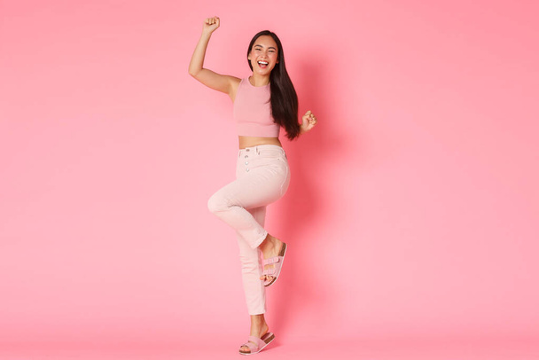 Mode, schoonheid en lifestyle concept. Succesvolle en winnende Aziatische vrouw in stijlvolle outfit, handen omhoog vrolijk, verheugen als feestelijk begin van de vakantie, gaan op vakantie, roze achtergrond - Foto, afbeelding