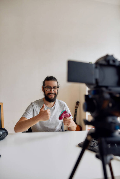 Male technology blogger in glasses showing thumbs up, holding game controller joystick while recording video blog or vlog about new gadgets at home - Fotó, kép