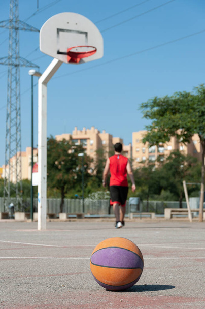 A vertical selective focus shot of a basketball in a court with the male player walking near the ring - Фото, изображение