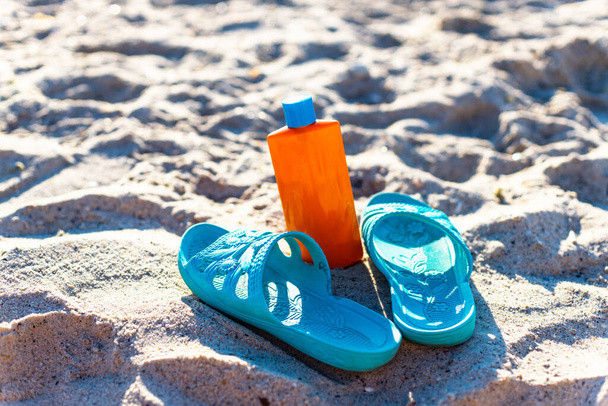 Kék gumi strand cipő és narancssárga műanyag fényvédő vagy krém palack a homokban a tenger közelében - Fotó, kép