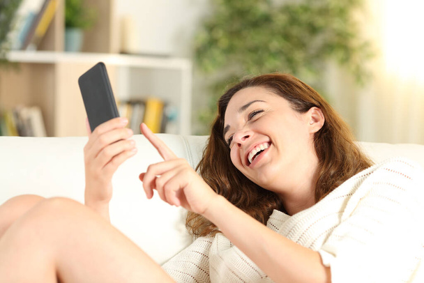 Happy girl laughing checking smart phone sitting on a sofa in the living room at home - 写真・画像
