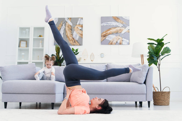 Young mom making stretching exercises and practicing yoga with baby girl at home. Health care and sports concept. - 写真・画像