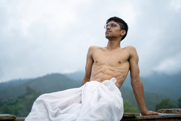 junger zerfetzter Mann sitzt in einem Dhoti und einer Brille mit verschwommenen Bergen im Hintergrund. - Foto, Bild