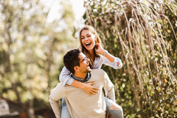 Usmívající se krásný mladý pár v lásce baví v parku. Pohledný muž dává prasátko zpět své přítelkyni. - Fotografie, Obrázek
