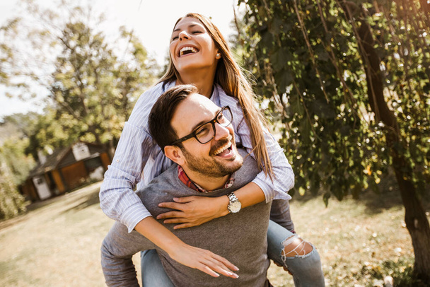Usmívající se krásný mladý pár v lásce baví v parku. Pohledný muž dává prasátko zpět své přítelkyni. - Fotografie, Obrázek