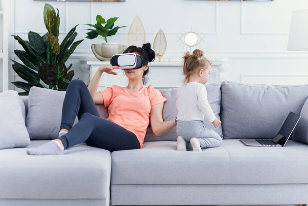 Pretty woman uses virtual reality goggles at home on sofa while her daughter watches cartoons. Technologies of the future. - Photo, Image