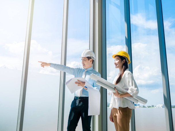 Asian architect Male and female inspecting construction work inside high-rise building near huge window glass on blue sky background. Couple engineer in hard hats holding blueprint in new building. - Photo, Image