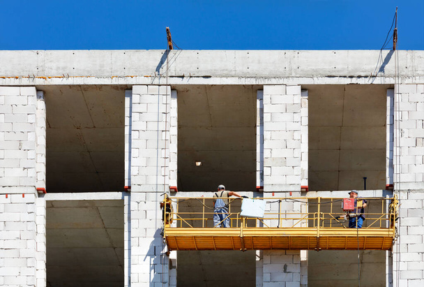 İnşaat beşiği inşaat halindeki bir evin duvarında işçiler varken, askıya alınan elektrik platformu yükseklikte güvenli bir iş için güvenilirdir.. - Fotoğraf, Görsel