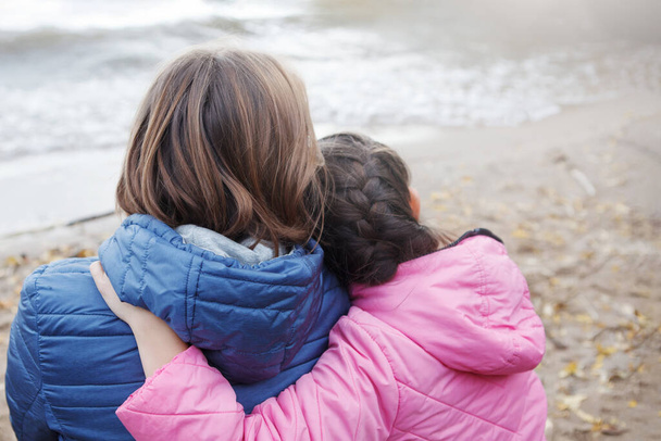 Mom and daughter sit hugging on the shore. Pink and blue jackets, long hair. Family happiness. Back view. Selective focus, blur background. - Photo, Image