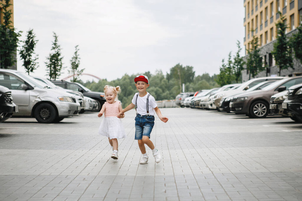 Junge und Mädchen laufen die Straße entlang. Eine Blondine mit Pferdeschwanz im rosa Kleid und ein Typ mit Baseballkappe zwischen Autos und Gebäuden. - Foto, Bild